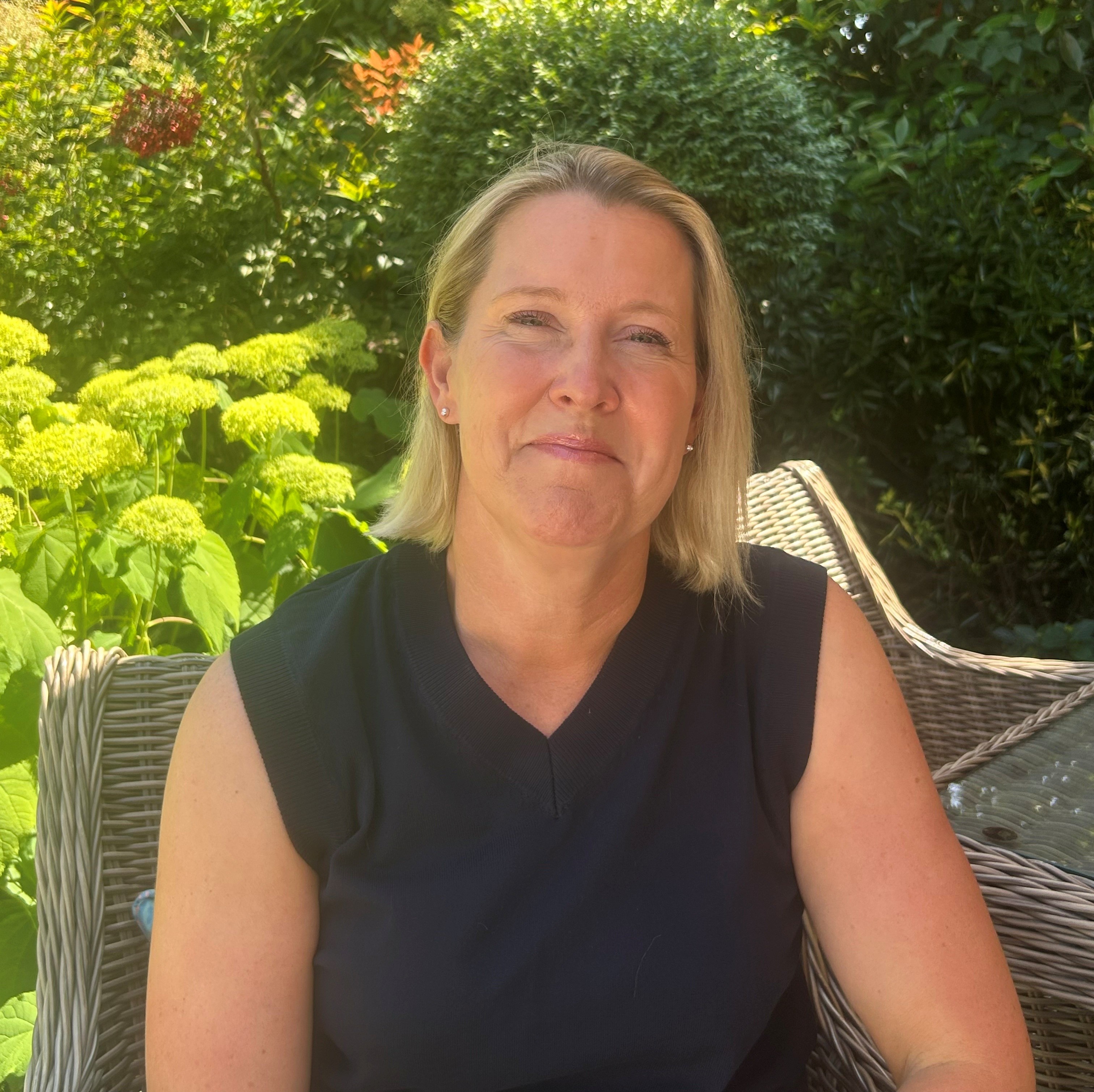 Madeleine Williams facing the camera and smiling whilst sitting outside in a sunny garden. Greenery features in the background.
